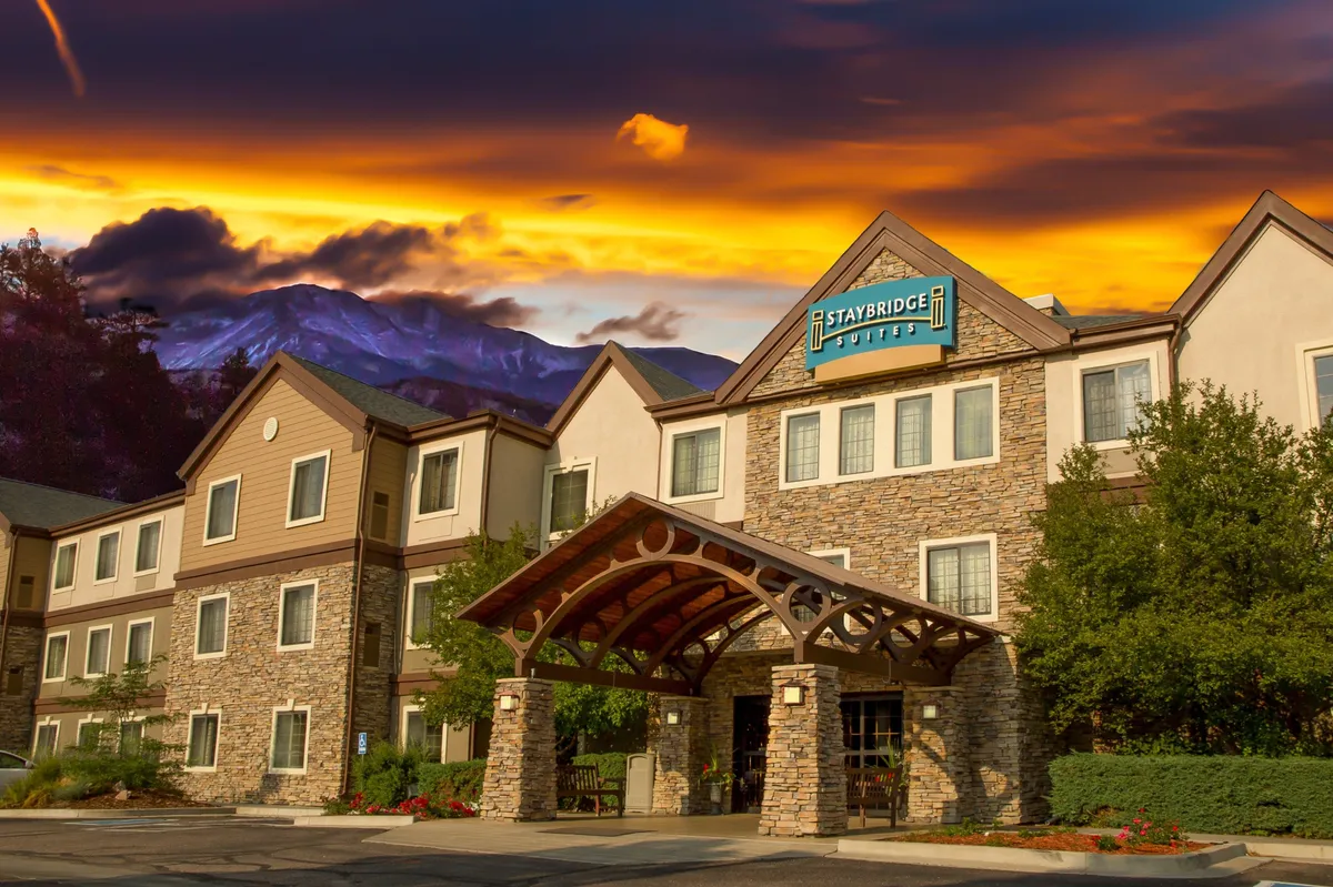 This image captures the welcoming entrance to the property, featuring a stone facade, a prominent hotel sign, and an inviting awning. The ambient light of sunset bathes the area, creating a warm, approachable atmosphere. Guests can be seen relaxing outside, enjoying the evening and the stunning mountain backdrop.