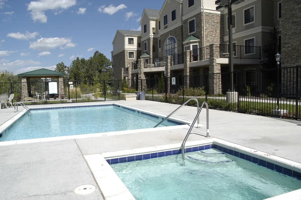 This exterior image centers on a sparkling swimming pool and adjacent hot tub, with the building’s facade and lush greenery creating a welcoming backdrop. Guests are captured enjoying relaxing moments in the hot tub and pool area, suggesting both leisure and opportunities for casual socializing. The environment is sunlit, reinforcing a resort-like, communal atmosphere.