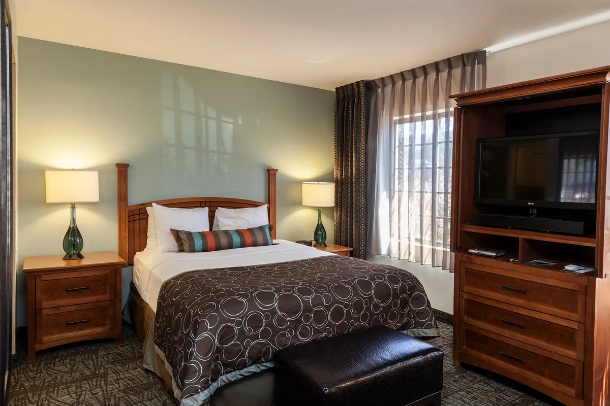 A comfortable interior space featuring a plush bed flanked by nightstands and lamps, with a television situated across from the bed and a window offering natural light. The ambiance is inviting and restful, suitable for relaxing or watching TV.