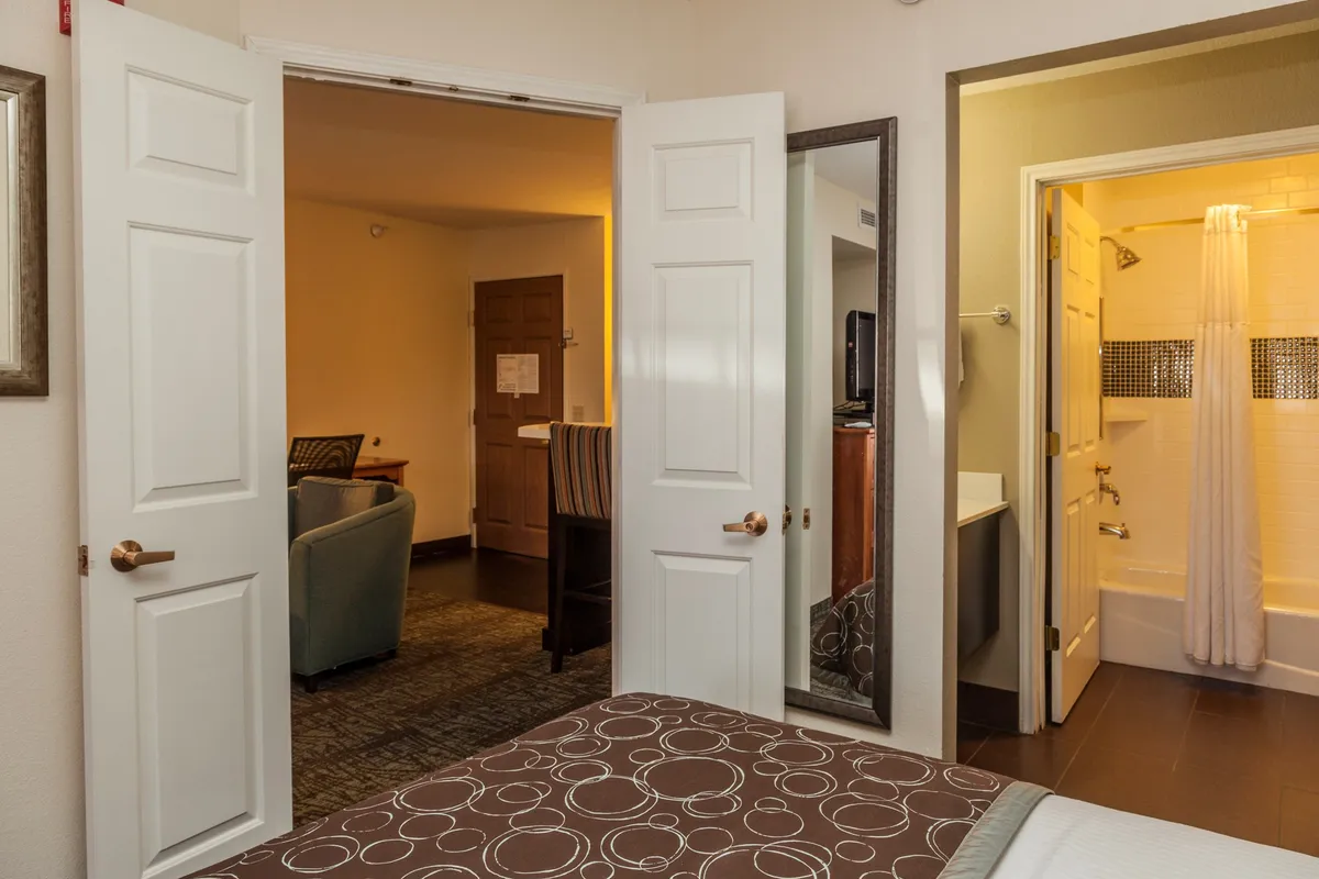 A bright, inviting room with a patterned bed in the foreground. Open doors connect the bedroom to a visible living area, and a mirror reflects light, making the space feel open and connected. The scene suggests easy transitions between lounging, socializing, and relaxing.