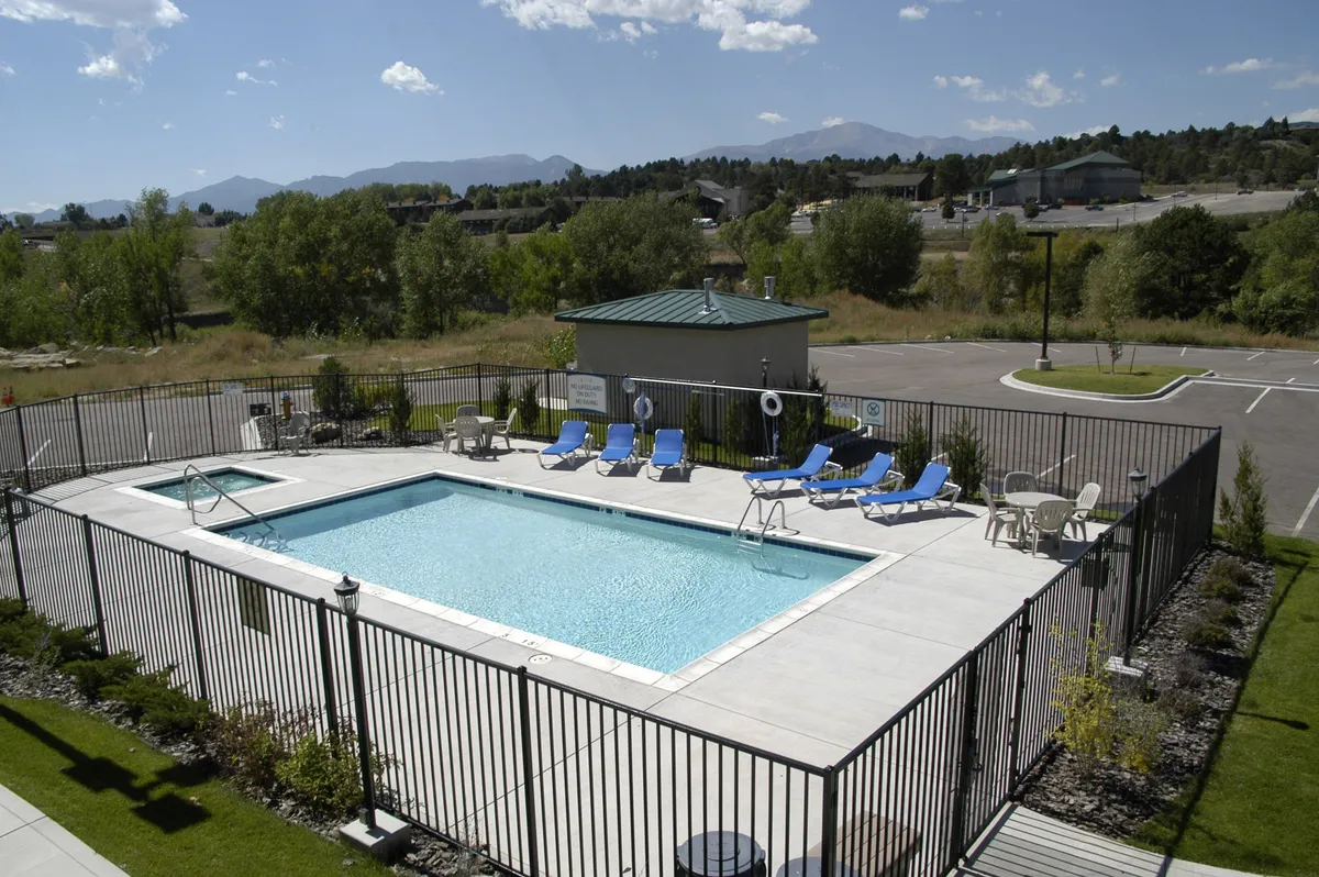 This exterior shot spotlights the resort-style swimming pool surrounded by lounge chairs, a gazebo, and lush green landscaping. The pool glistens under the daylight, with reflections hinting at both morning and sunset use. People are seen relaxing and socializing poolside, enjoying leisure time in a refreshing setting.